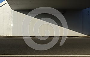 White concrete wall under an underpass divided in two obliquely by a shadow. Asphalt road in front.