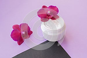 White container with cream for face and body with two magenta colored orchid flowers on purple and black background. Concept of de