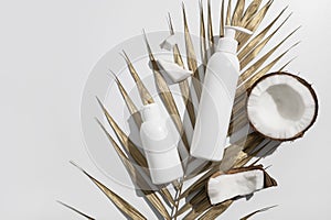 White cosmetic bottles, fresh coconut with sunny contrast shadows, gold palm leaf on gray background. Organic natural cosmetics