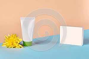White cosmetic tube with green cap and no label next to yellow chrysanthemum flower and box against colorful studio