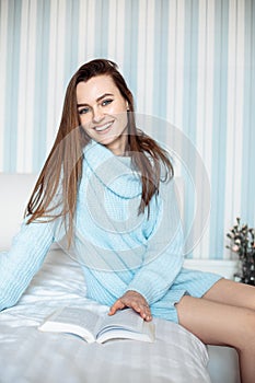 White cozy bed and beautiful young woman, sitting reading book, concepts of home and comfort, looking at camera