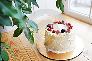 White cream cake decorated with berries on wooden background near the window