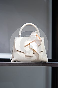 White cream handbag for women with cream ribbon tied on the handle of the bag is placed on a wooden shelf