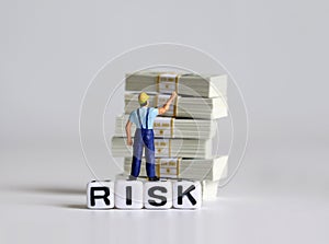 A white cube with `RISK` text. Bundle of $100 bills and a miniature man.