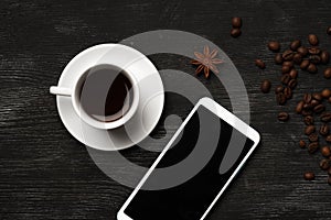 White cup with anise coffee and a mobile phone with a blank screen on a black background with coffee beans and place for text top