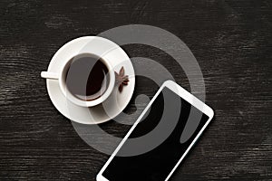 White cup with anise coffee and a mobile phone with a blank screen on a black background with place for text top view