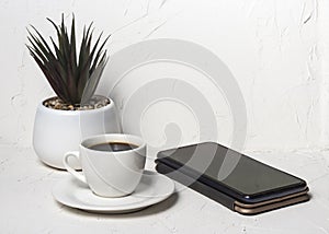 White cup with black aromatic coffee on a white abstract background with a potted flower in the background with a smartphone