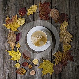 White cup with black coffee in a circle of colored dry leaves on old wooden background. Leaves in a circle with mug of