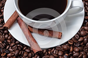 White cup of black coffee with plate and three cinnamon sticks on beans background half view macro