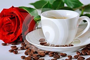 White cup of black coffee with a red rose and coffee beans scattered on a white table
