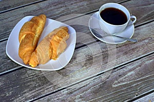 A white cup of black coffee on a saucer with a spoon, next to a plate with two fresh croissants