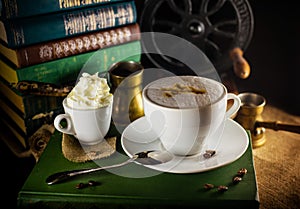 White cup with cappuccino and one else small with cream stand on book. Atmosphere of vintage library with old books and grinder on