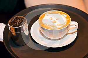 White cup of cappuccino with a picture of a bear`s face, on a dark tray in the hand of an offspring close-up