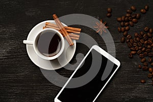 White cup with cinnamon and anise coffee sticks and a mobile phone with a blank screen on a black background with coffee beans and