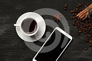 White cup with cinnamon and anise coffee sticks and a mobile phone with a blank screen on a black background with coffee beans and