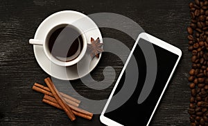 White cup with cinnamon and anise coffee sticks and a mobile phone with a blank screen on a black background with coffee beans and