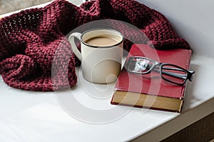 White cup with coffee, book, glasses and burgundy knitted scarf by the window