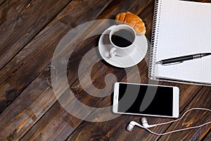 White cup of coffee with headphones and mobile phone and lying next to notebook with pen, croissant