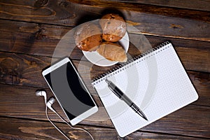 White cup of coffee with headphones and mobile phone and lying next to notebook with pen, croissant