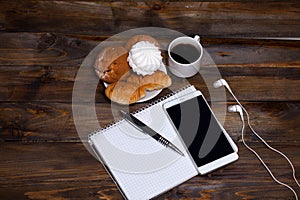 White cup of coffee with headphones and mobile phone, and lying next to notebook with pen, croissant, marshmallow