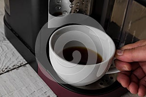 A white cup of coffee with prepaired drink still standing at the coffee machine platform. Breakfast and home equipment concept.