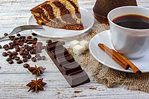 A white cup with coffee on sacking and a piece of amazing chocolate cake on a plate with a fork. A chocolate bar, coffee beans, an