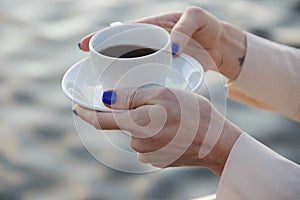 White Cup of coffee with a saucer in the graceful hands of a woman in beige clothes