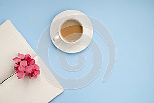 White cup of coffee silver notepad, pink flower on a blue background. Flat lay. copy space. top view