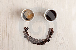 White cup of coffee and smile made of roasted coffee beans on light coloured wooden table