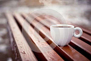 A white cup of coffee stands on wooden boards in natural environment in the winter.