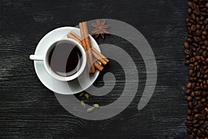 White cup with coffee sticks of cinnamon green cardamom and anise on a black background with coffee beans and place for text top