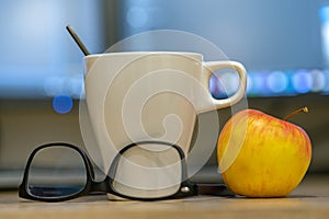 White cup of coffee or tea, glasses and a red apple on blurred background of personal computer screen