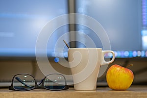 White cup of coffee or tea, glasses and a red apple on blurred background of personal computer screen
