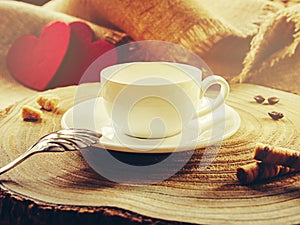 White cup with coffee, on a wooden background in the morning sun. Two red hearts in the background