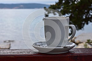 White cup of coffee at wooden bench/ sea and nature as background