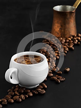 White cup with espresso on a black background, steam rises above the cup. Roasted coffee beans are located around a cup of coffee