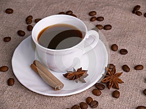 A white cup of espresso stands on a burlap table with scattered coffee beans, a cinnamon stick and stars of anise