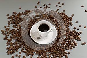 White cup with espresso on a white saucer among coffee beans on a gray background.