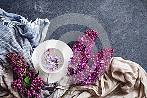 White cup with flowers on a saucer, a bouquet of lilac flowers and a blue drape striped scarf on a dark background