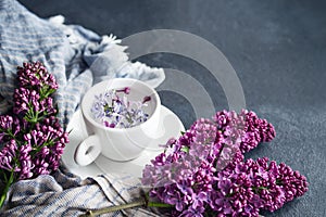 White cup with flowers on a saucer, a bouquet of pink lilac flowers and a blue drape striped scarf