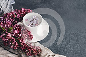 White cup with flowers on a saucer, a bouquet of pink lilac flowers and a blue drape striped scarf