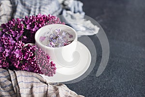 White cup with flowers on a saucer, a bouquet of pink lilac flowers and a blue drape striped scarf