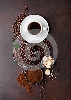 White cup of fresh raw organic coffee with beans and ground powder with cane sugar cubes with coffee tree leaf on brown background