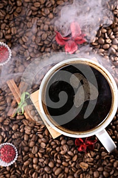 White cup full of coffee beans on a coffee beans background with red chocolate candy and red flowers. Morning espresso. Coffee mug