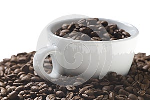 White Cup full of coffee beans on roasted coffee beans background.