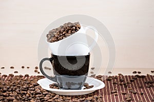 White cup full of roasted coffee beans standing on a black on white plate standing on tablemat.