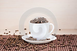 White cup full of roasted coffee beans standing on a white plate on tablemat.