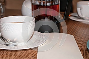 White cup, glass kettle with dark tea and empty paper blank with copy space on wooden table