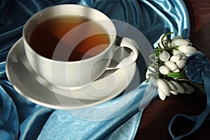 A white cup with green tea on a saucer and a small bouquet of spring white snowdrops, tied with a ribbon. Rustic still