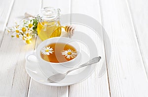 White cup of herbal tea with chamomile flowers and honey on the white wooden background. Natural alternative medicine